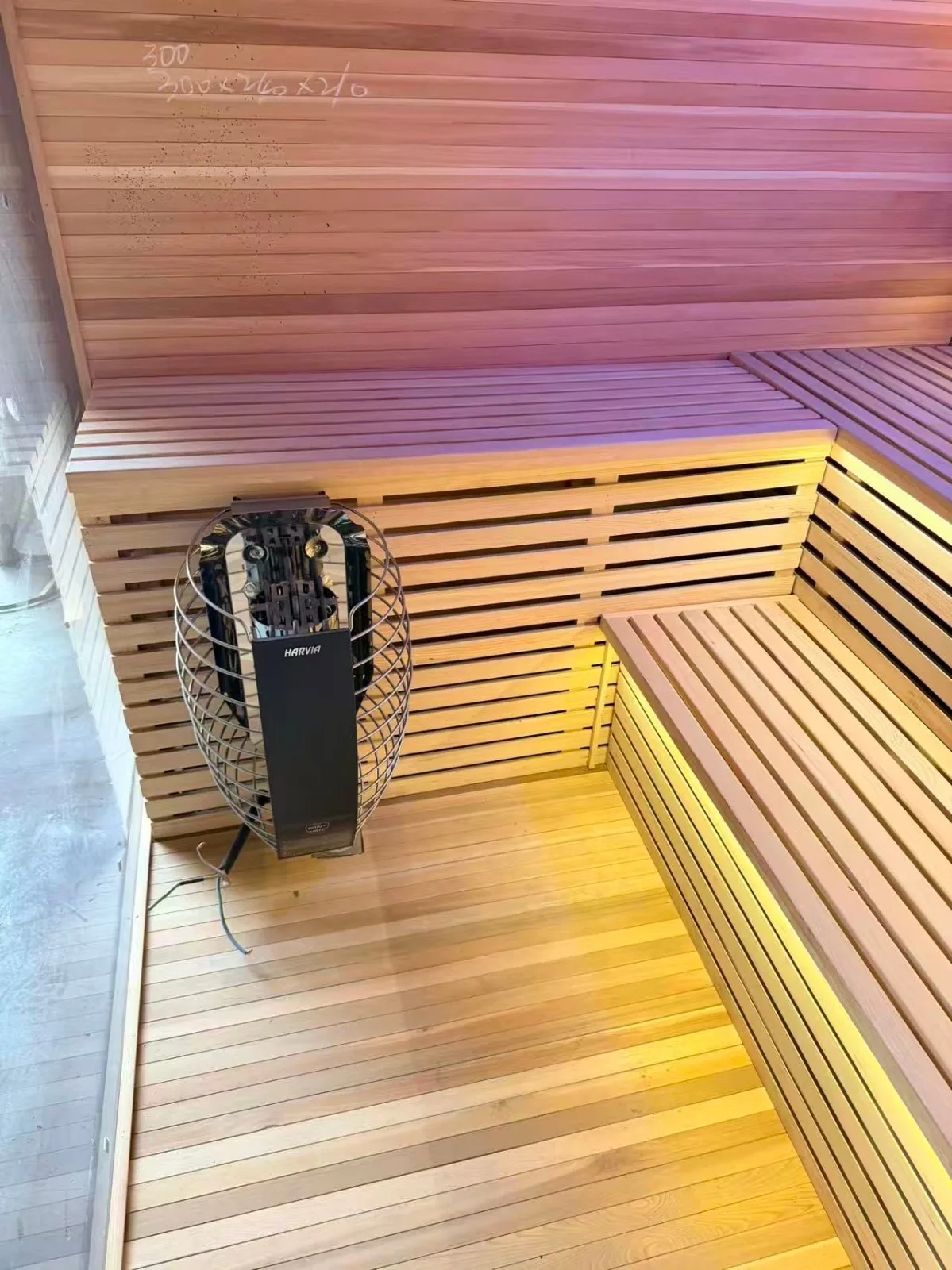 Indoor sauna corner during dry-fit: wall-mounted Harvia cage heater, L-bench in light timber, duckboard floor